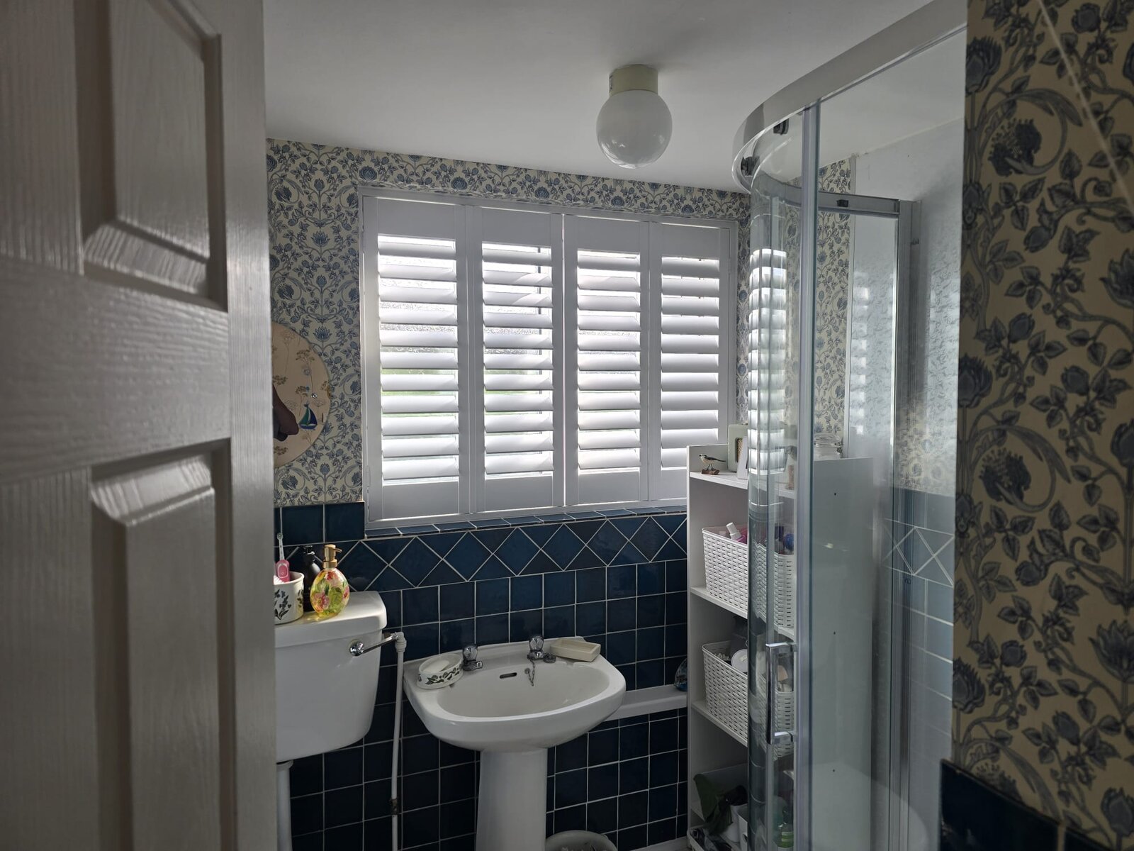White hardwood full-height shutters on a bathroom window in a Chacombe home with blue tiles and floral wallpaper