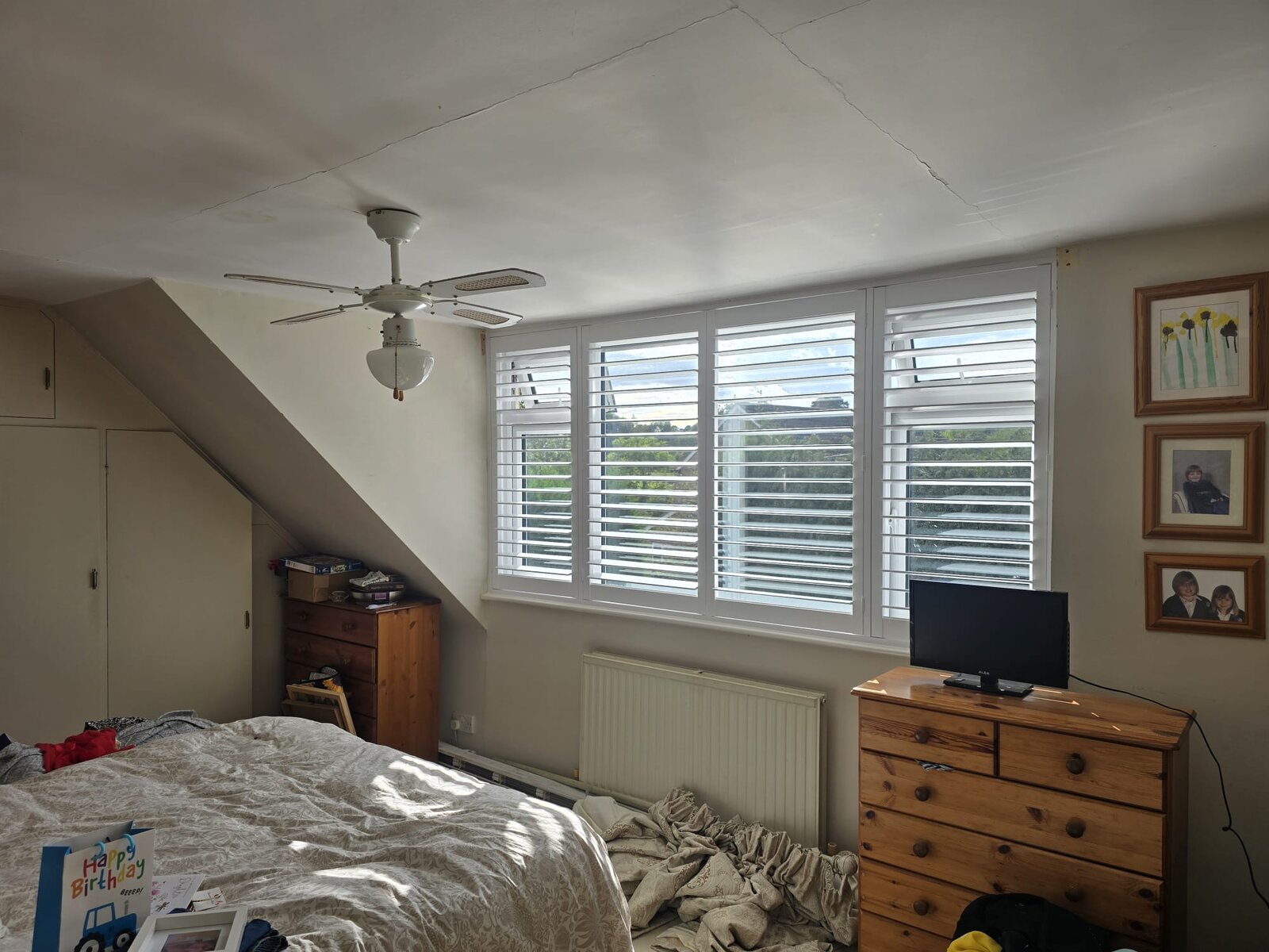 Four-panel white hardwood full-height shutters on a bedroom window in Chacombe with a sloped ceiling