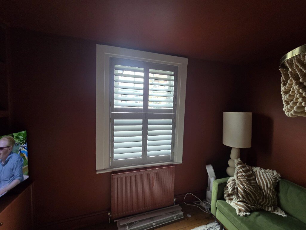 Slate Grey hardwood tier-on-tier shutters with 89mm louvres open on a Croydon living-room window beside a green sofa