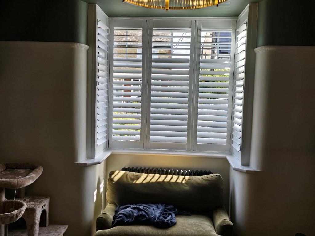 Three-sided bay with Snow White full-height hardwood shutters casting louvre shadows on a green armchair