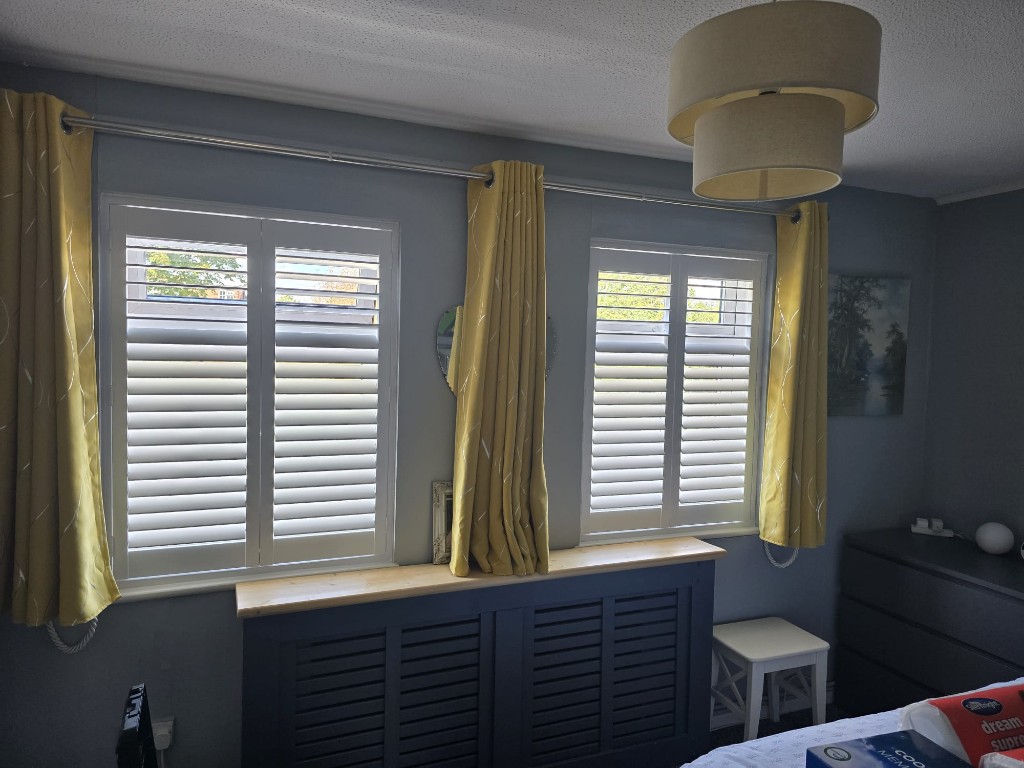 Twin Snow White full-height hardwood shutters in a bedroom with mustard curtains — top louvres open, lower closed
